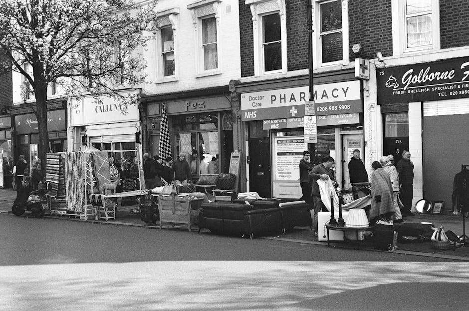 A street scene featuring a variety of household furniture and large items arranged along the pavement in front of commercial buildings, including a black upholstered sofa, a white appliance, and vario